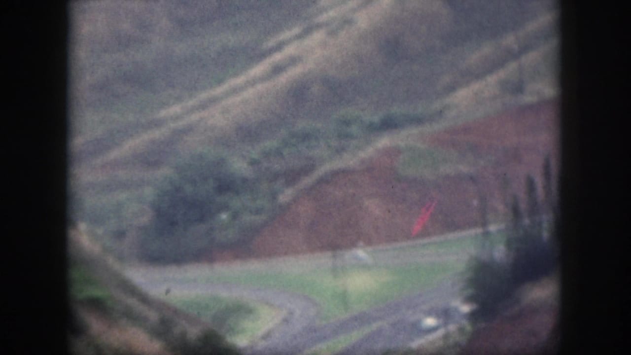 1959 Oahu home-movie view of cars on a curving road below muted hills, trees, and exposed reddish soil.