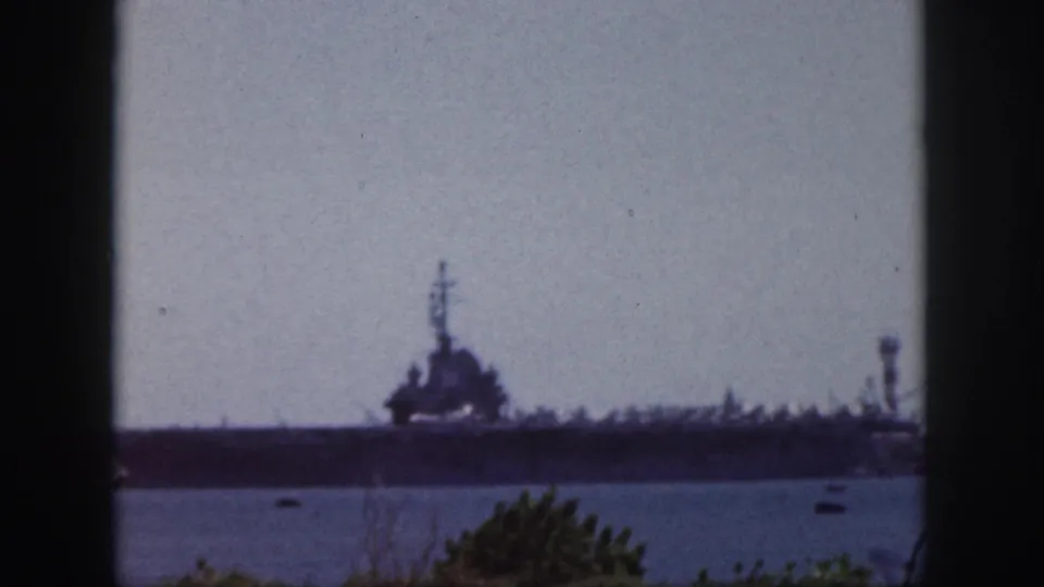 Distant large naval vessel in Oahu harbor, seen through dark window framing on faded 8mm color film.
