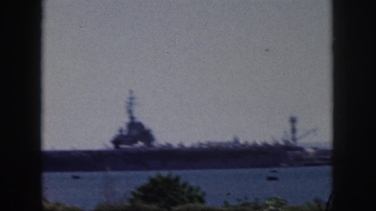 1959 Oahu home-movie frame showing a large naval vessel across harbor water, with small boats and foreground foliage.