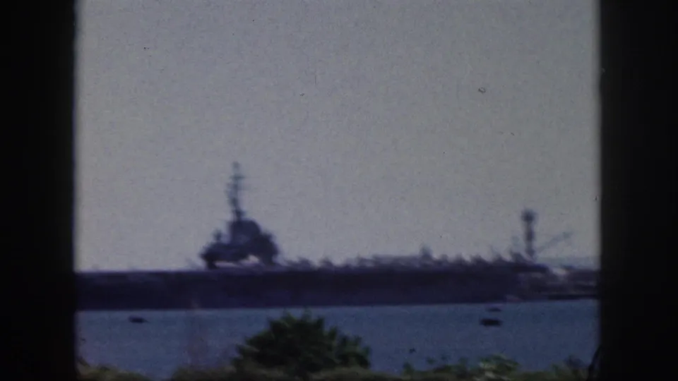 1959 Oahu home-movie frame showing a large naval vessel across harbor water, with small boats and foreground foliage.