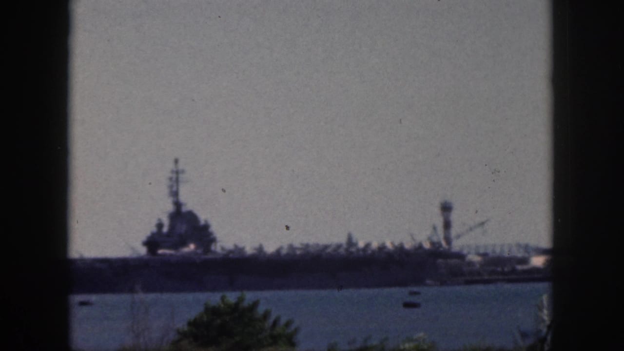 1959 Oahu 8mm frame showing a distant large military ship across harbor water, with haze, vegetation, and dark foreground edges.