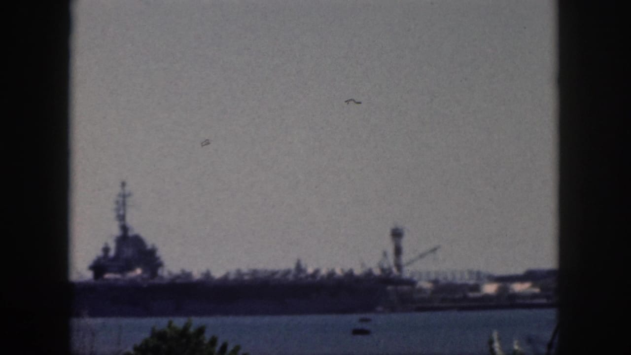 Distant naval vessel and harbor structures beyond calm water.