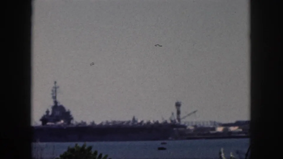 Distant naval vessel in an Oahu harbor, with industrial structures and small boats visible through soft 8mm color grain.