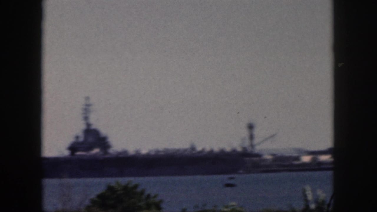 Soft, slightly blurred 1959 Oahu harbor frame with a large ship, tower-like structure, blue water, and foreground vegetation.