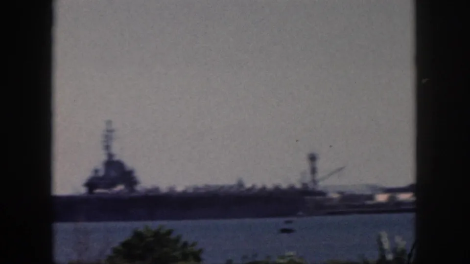 Soft, slightly blurred 1959 Oahu harbor frame with a large ship, tower-like structure, blue water, and foreground vegetation.
