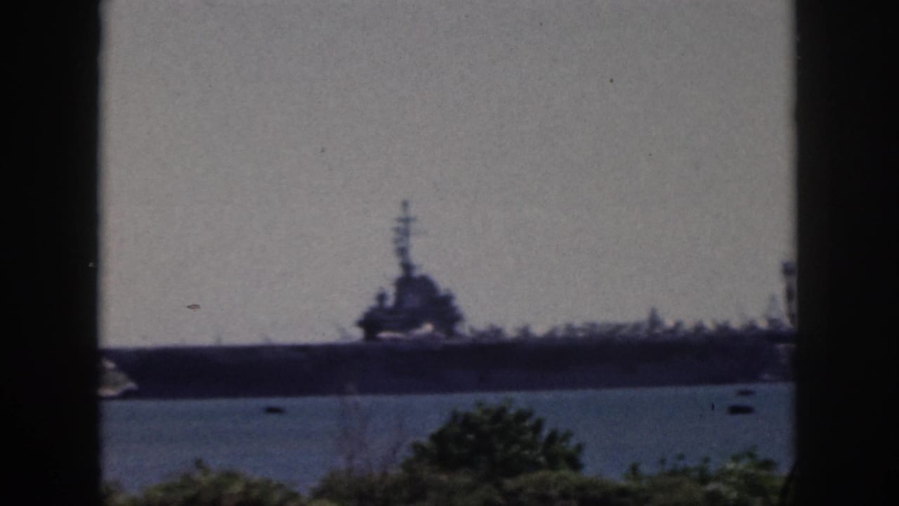 1959 Oahu home-movie frame showing a distant large naval ship across water, partly framed by dark vertical window bars.