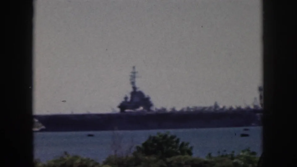 1959 Oahu home-movie frame showing a distant large naval ship across water, partly framed by dark vertical window bars.