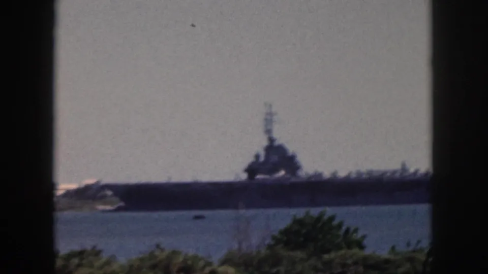 Distant naval vessel off Oahu in 1959, seen across blue water with a dark window frame and green foliage in the foreground.
