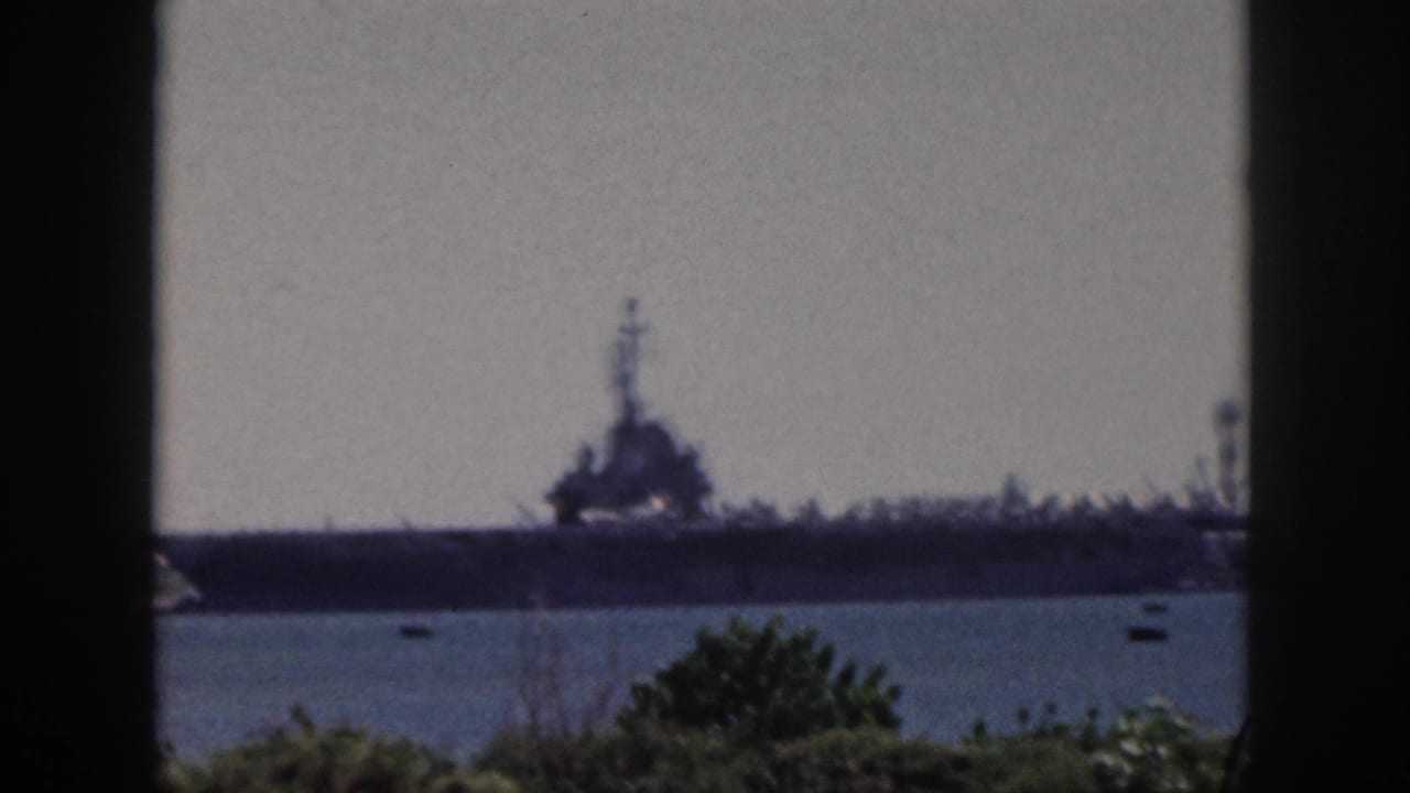 1959 Oahu footage frame with a large naval ship across the water, framed by a dark window edge and low green foliage.
