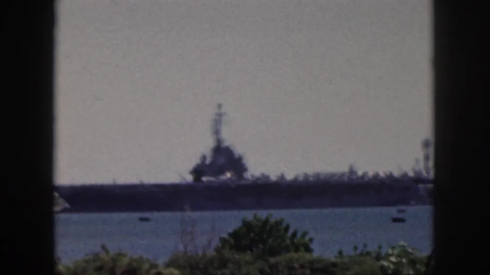 1959 Oahu footage frame with a large naval ship across the water, framed by a dark window edge and low green foliage.