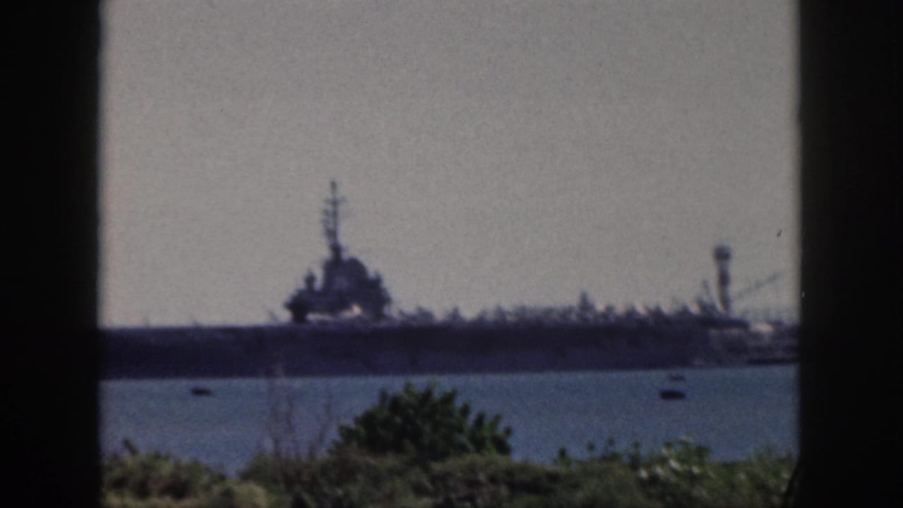 1959 Oahu home-movie frame showing a distant naval aircraft carrier across harbor water, partially bordered by a dark window frame.