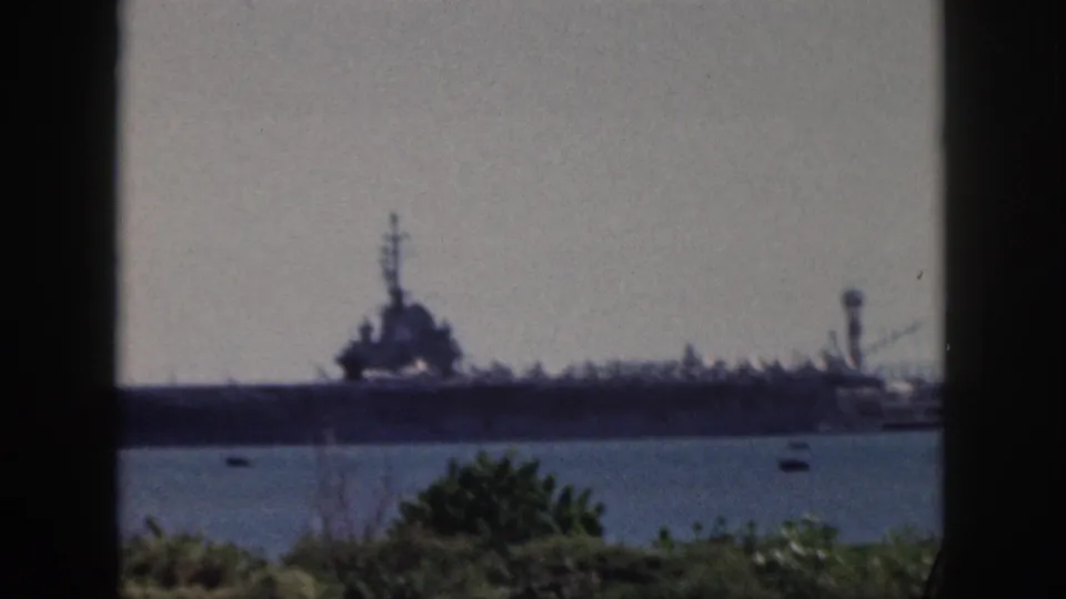 1959 Oahu home-movie frame showing a distant naval aircraft carrier across harbor water, partially bordered by a dark window frame.