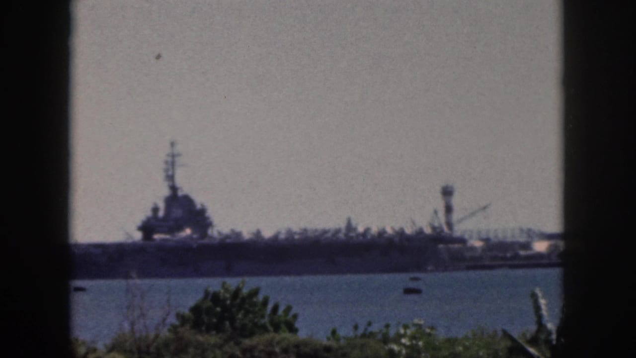 1959 Oahu harbor view with a naval aircraft carrier in the distance, small boats on the water, and foliage along the lower edge.