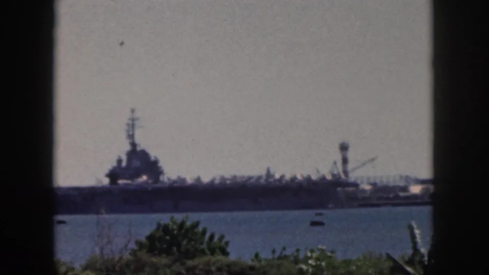 1959 Oahu harbor view with a naval aircraft carrier in the distance, small boats on the water, and foliage along the lower edge.
