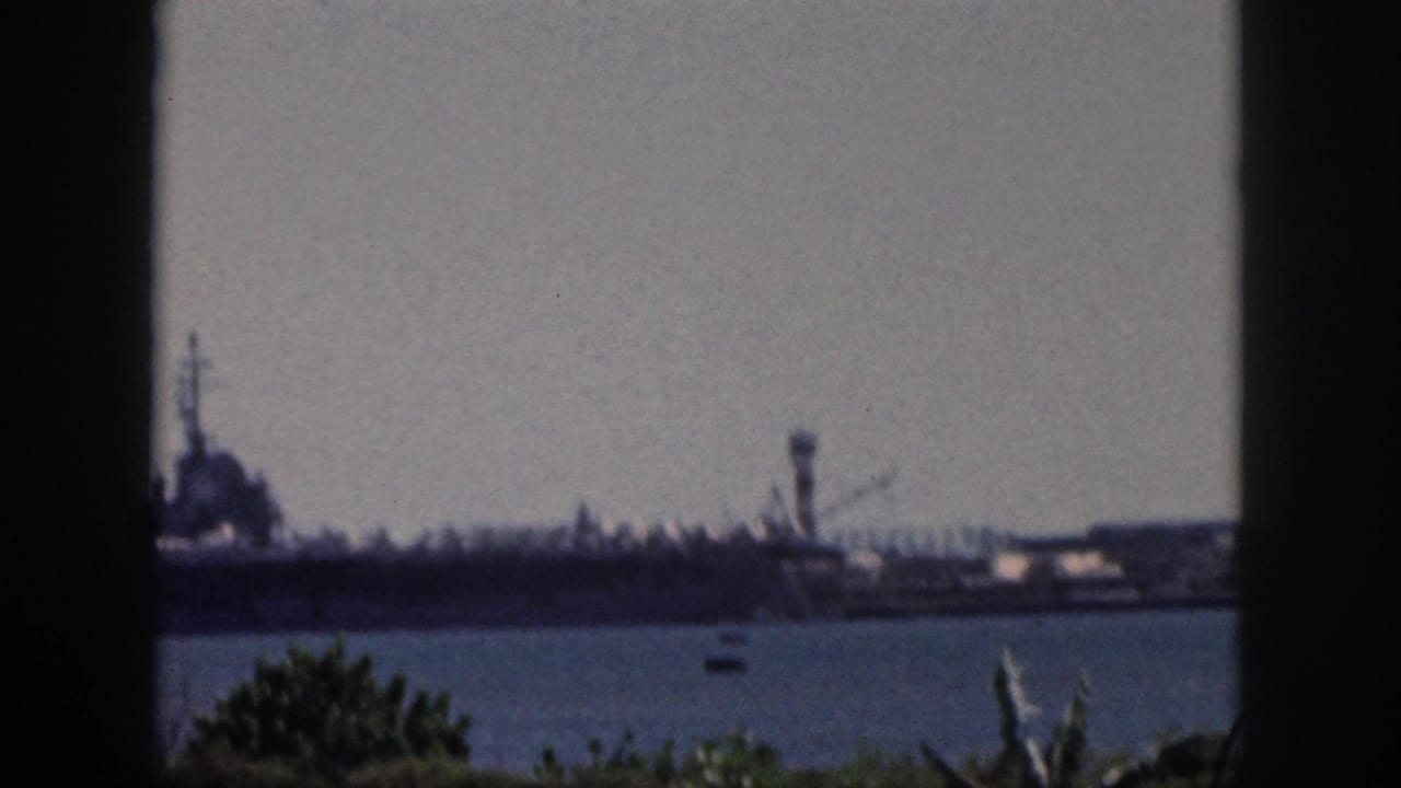 Soft 8mm frame from Oahu in 1959 showing the carrier partly cropped at left, with harbor water and plants in the foreground.