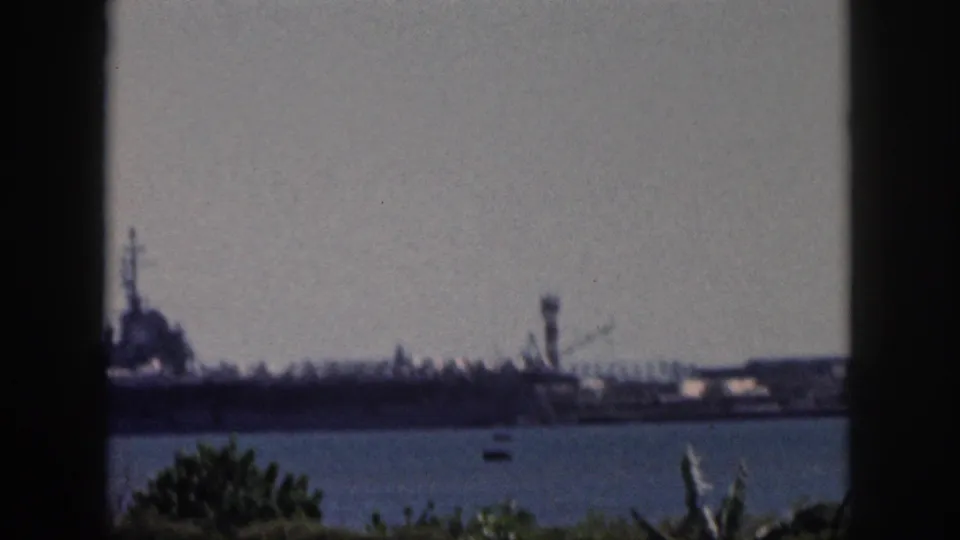 Soft 8mm frame from Oahu in 1959 showing the carrier partly cropped at left, with harbor water and plants in the foreground.