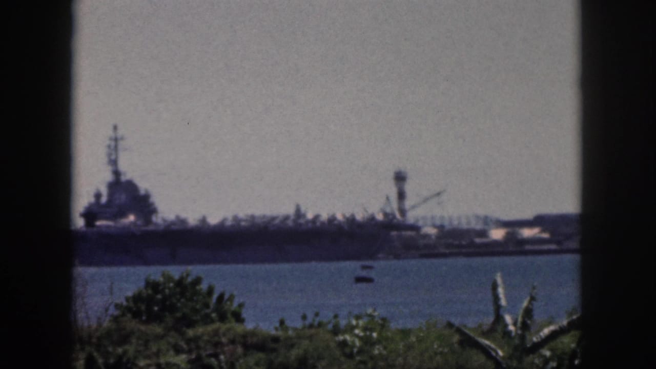 1959 Oahu 8mm frame showing a distant naval carrier across the harbor, with a dark window frame and shoreline plants in the foreground.