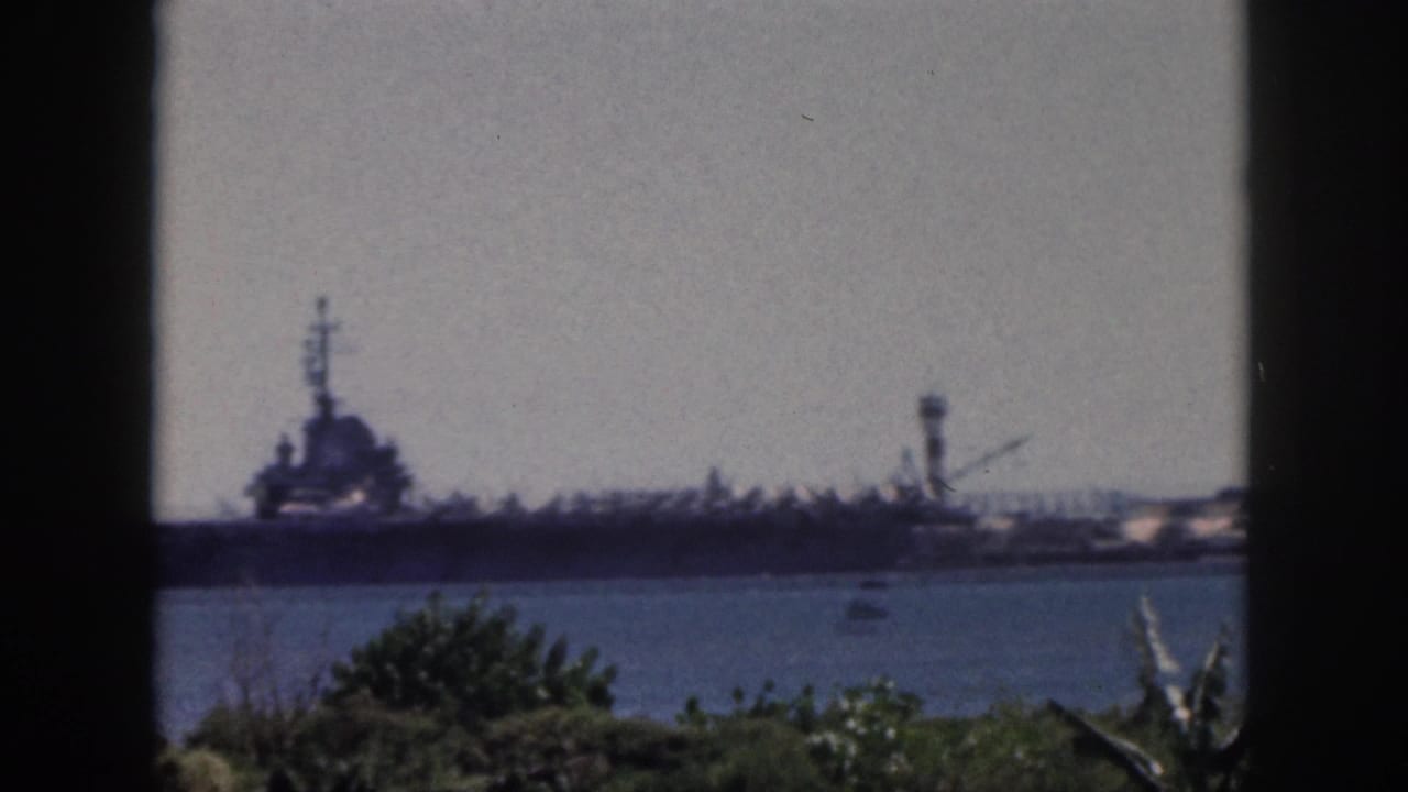 1959 Oahu home-movie frame showing a distant naval ship across calm harbor water with tropical foliage in the foreground.