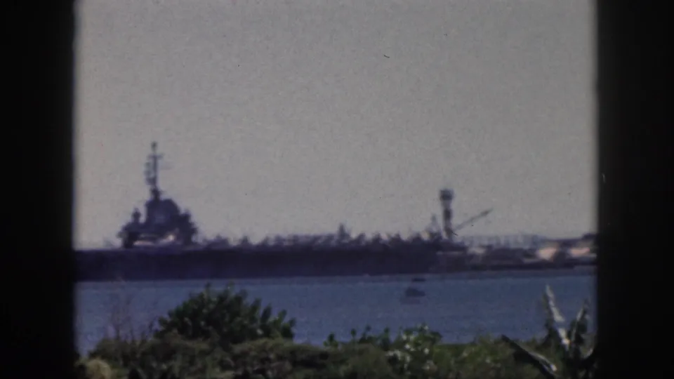 1959 Oahu home-movie frame showing a distant naval ship across calm harbor water with tropical foliage in the foreground.