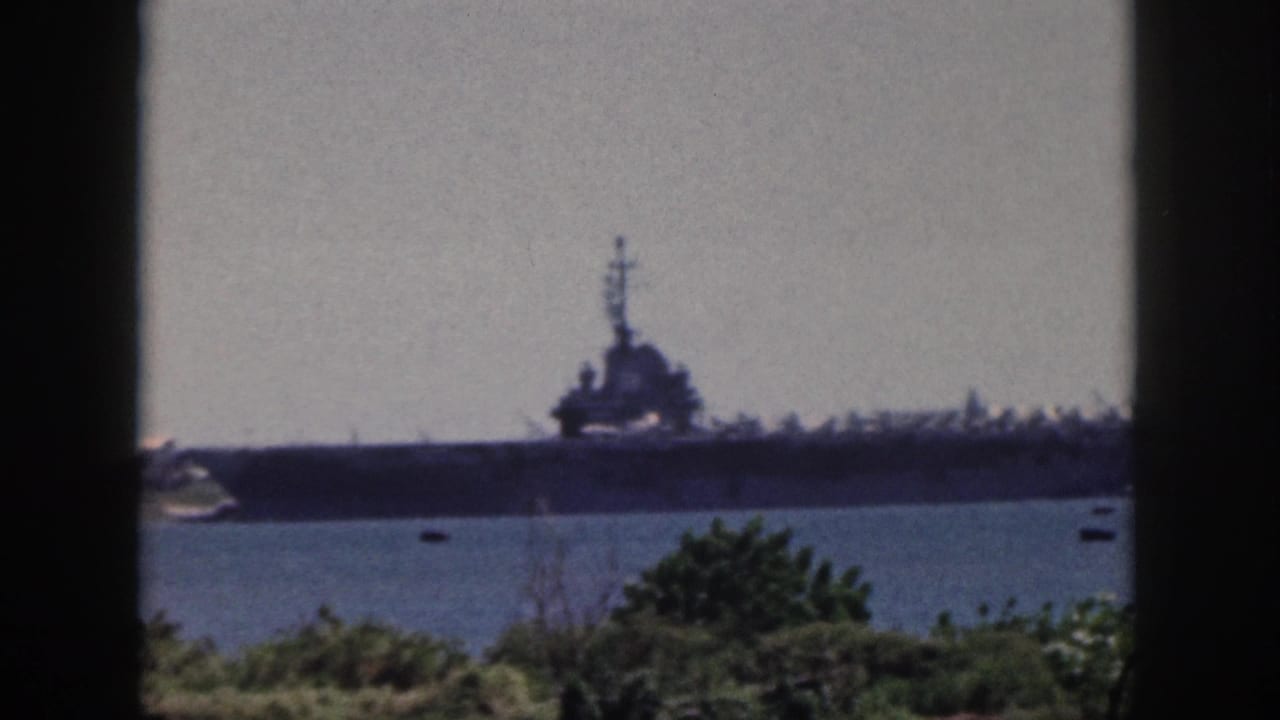 1959 Oahu home-movie frame showing a distant large naval ship across blue harbor water, partly framed by dark vertical bars and shoreline greenery.