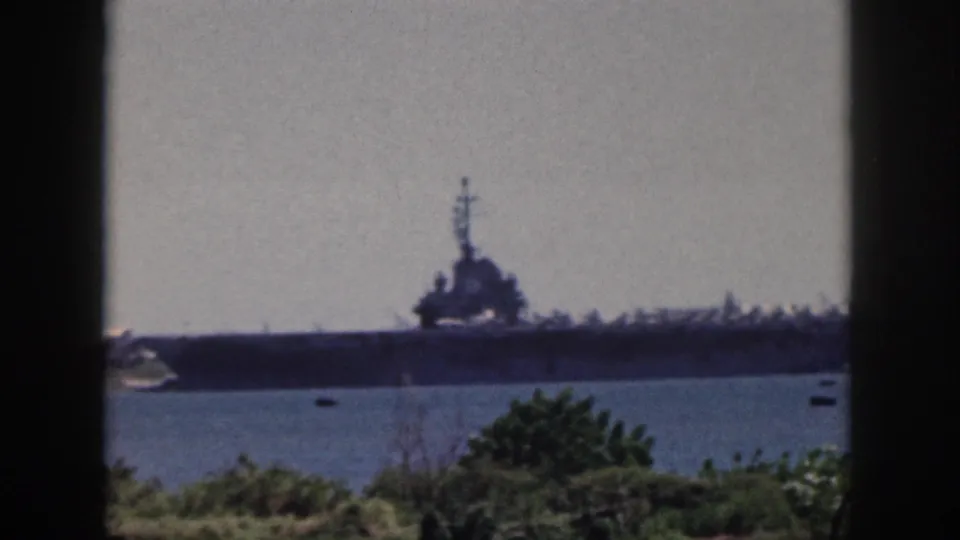 1959 Oahu home-movie frame showing a distant large naval ship across blue harbor water, partly framed by dark vertical bars and shoreline greenery.