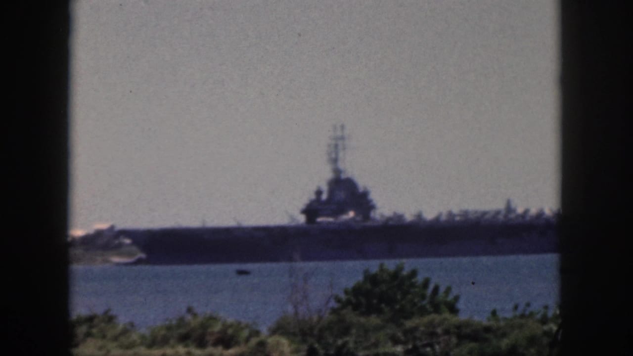 Oahu, 1959 footage frame with a large carrier-like naval vessel across the water, seen beyond greenery and dark window-like framing.