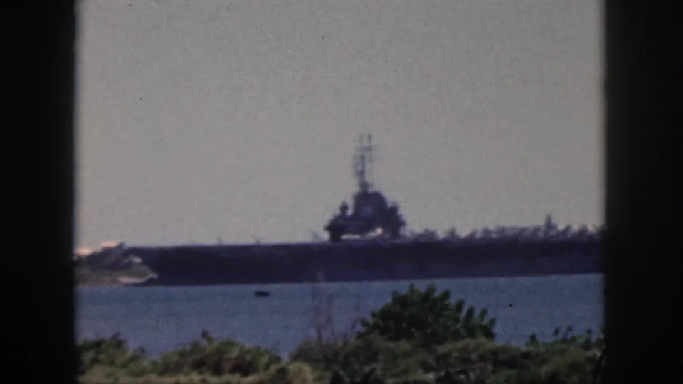 Oahu, 1959 footage frame with a large carrier-like naval vessel across the water, seen beyond greenery and dark window-like framing.