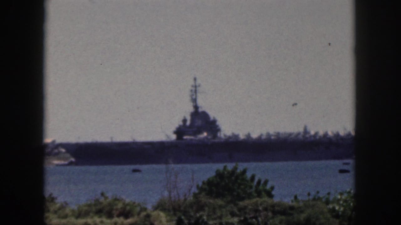 Soft, grainy 1959 Oahu home-movie view of a large naval ship docked or moored beyond harbor water, with shoreline vegetation in the foreground.