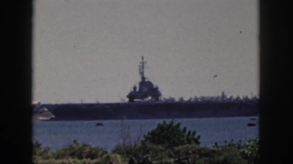Soft, grainy 1959 Oahu home-movie view of a large naval ship docked or moored beyond harbor water, with shoreline vegetation in the foreground.