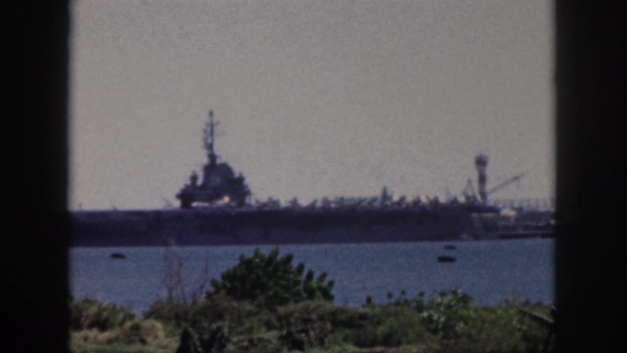 1959 Oahu home-movie frame showing a large naval ship across the water beyond shoreline vegetation.
