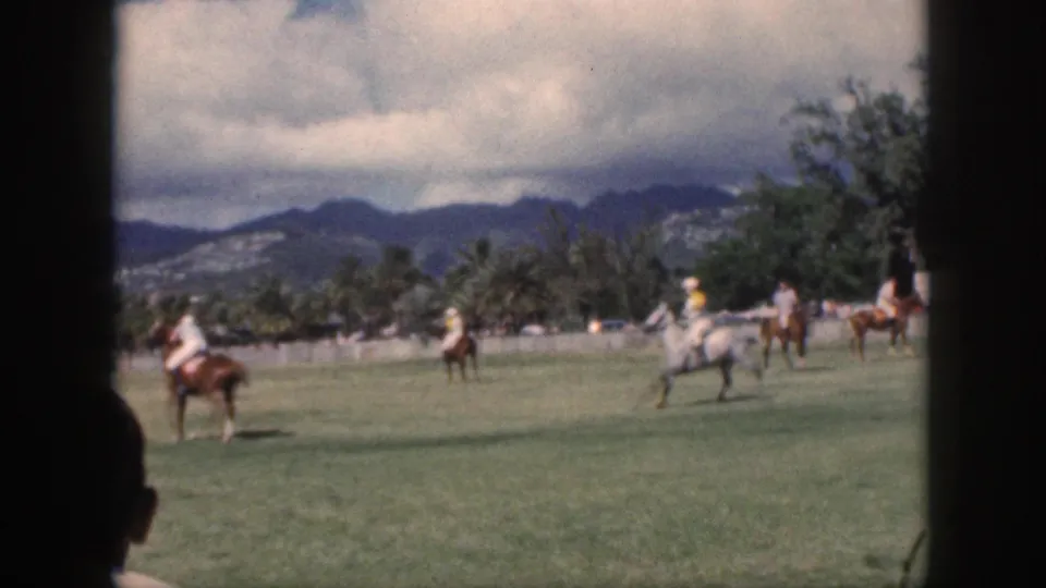 1959 Oahu home-movie frame of polo players on horseback spread across the field with mountains beyond.