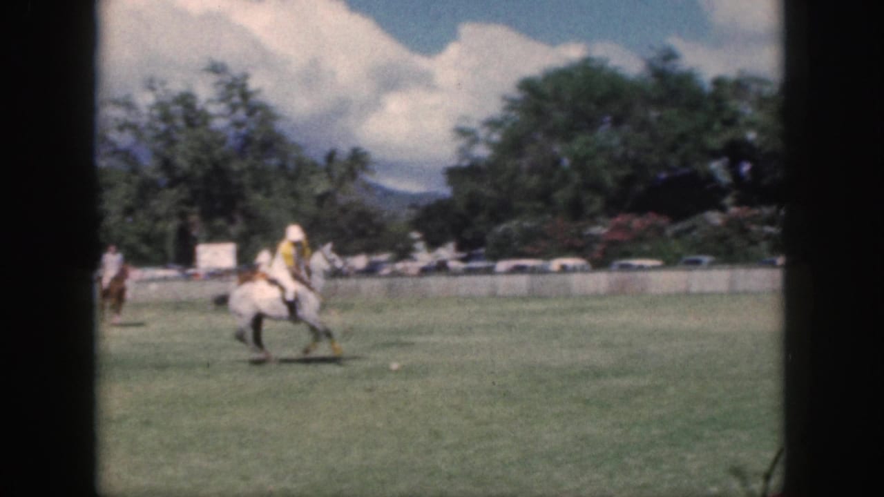 1959 Oahu home-movie frame of polo riders on a grassy field, with trees, parked cars, mountains, and clouds in the background.