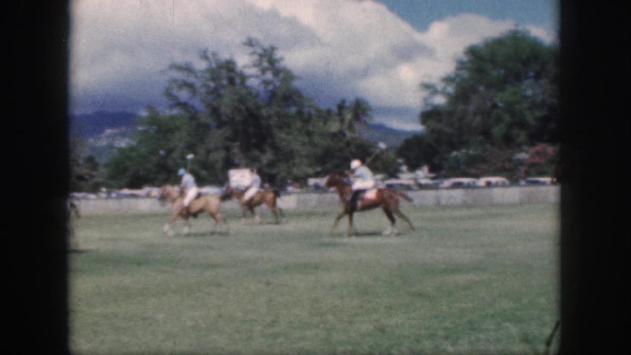 Distant Oahu polo riders cross the field in a 1959 8mm home-movie frame, backed by trees, mountains, and a bright cloudy sky.