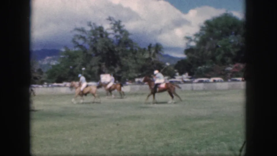 Distant Oahu polo riders cross the field in a 1959 8mm home-movie frame, backed by trees, mountains, and a bright cloudy sky.