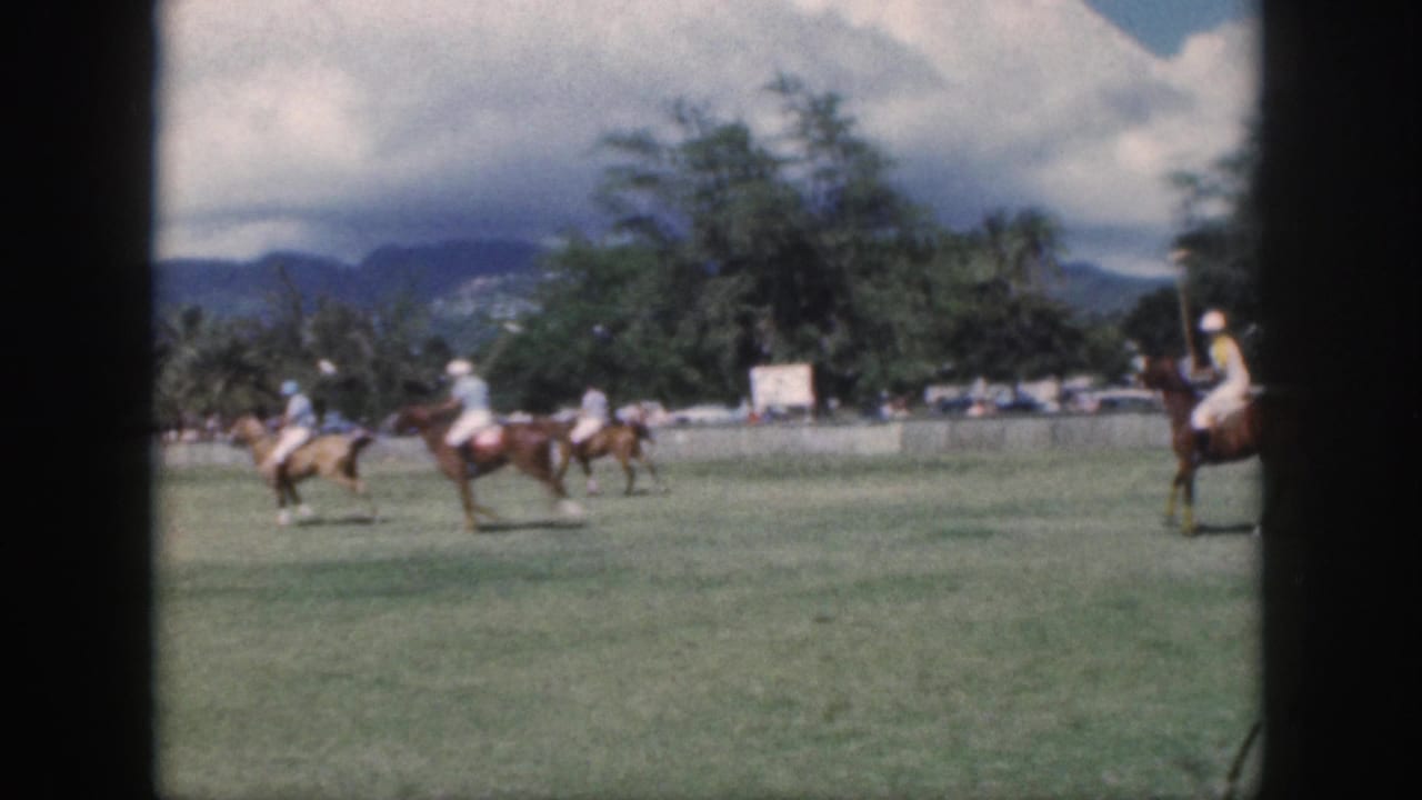 1959 Oahu home-movie frame showing mounted polo players crossing a grassy field beneath clouds and distant mountains.