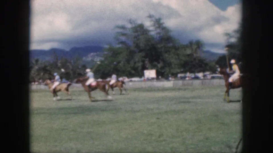 1959 Oahu home-movie frame showing mounted polo players crossing a grassy field beneath clouds and distant mountains.