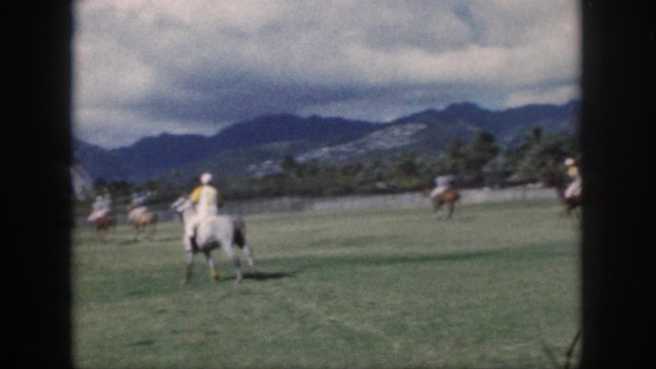 Oahu 1959 home-movie frame with a mounted polo player seen from behind on a grassy field below mountain ridges.