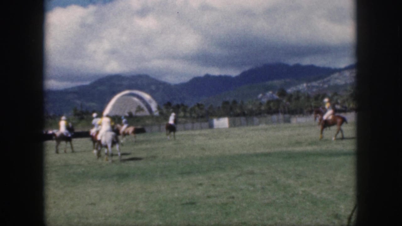 1959 Oahu polo scene with mounted players on a grassy field and a distant white arched structure against mountains.