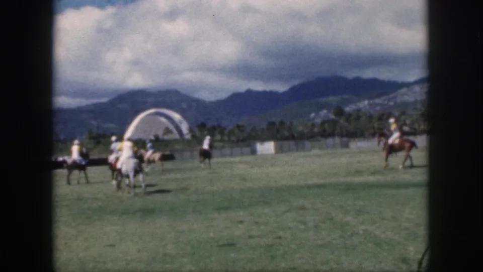1959 Oahu polo scene with mounted players on a grassy field and a distant white arched structure against mountains.