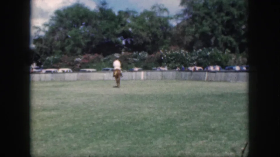The rider appears farther from the camera on the Oahu grass field, with a low wall, cars, and dense greenery behind.
