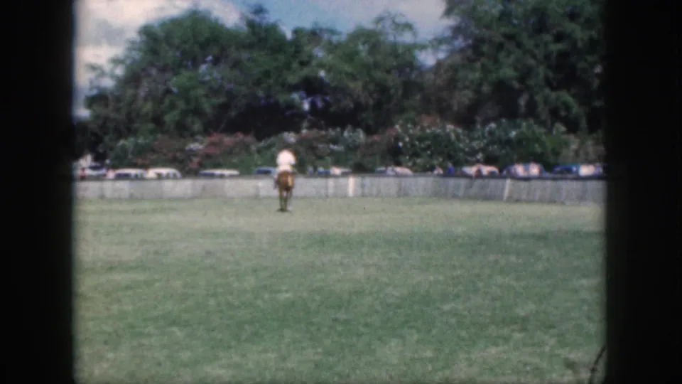 A horseback rider is seen from behind in the 1959 Oahu field, with vintage cars and leafy trees along the far edge.