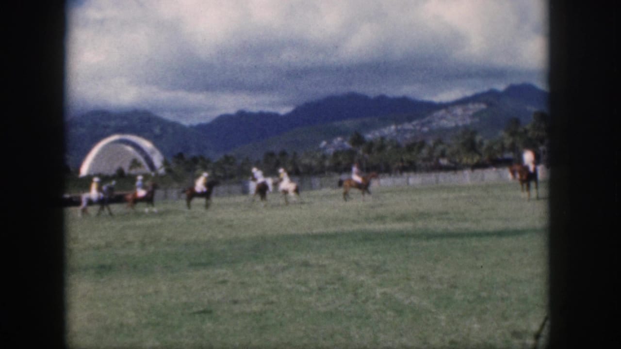 1959 Oahu home-movie frame showing riders in play near a white arched structure with palms and mountains in back.