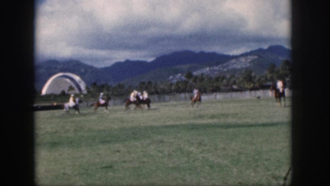 1959 Oahu home-movie frame showing polo players on horseback moving across a green field beneath cloudy mountains.