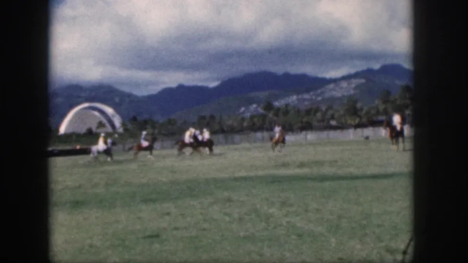 1959 Oahu home-movie frame showing polo players on horseback moving across a green field beneath cloudy mountains.
