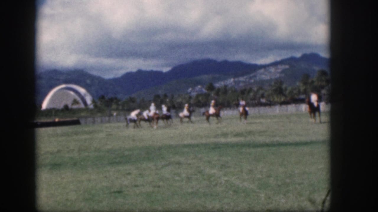 Mounted polo players cluster on the field with a white arch, palms, and mountains in the distance.
