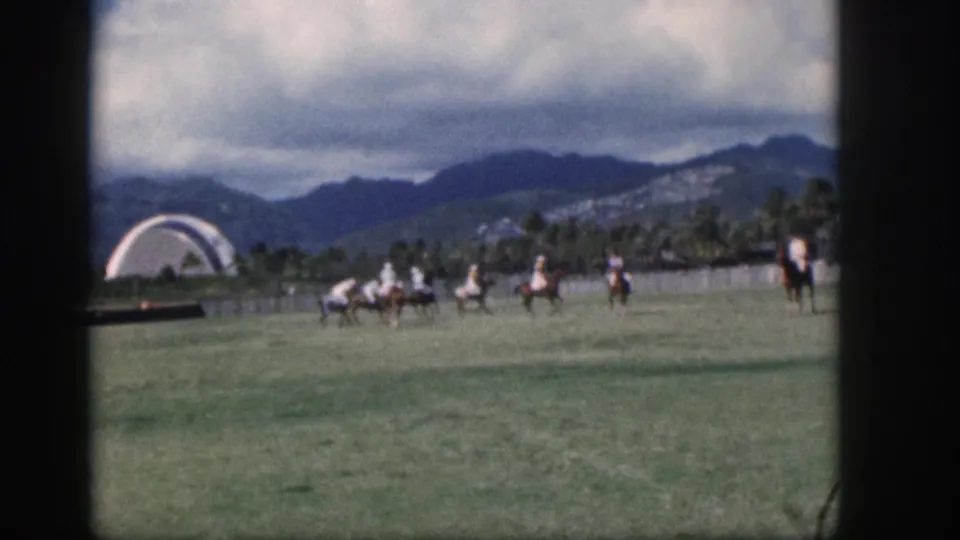 1959 Oahu 8mm frame of polo riders grouped on a grass field, with palms and mountains behind the play.
