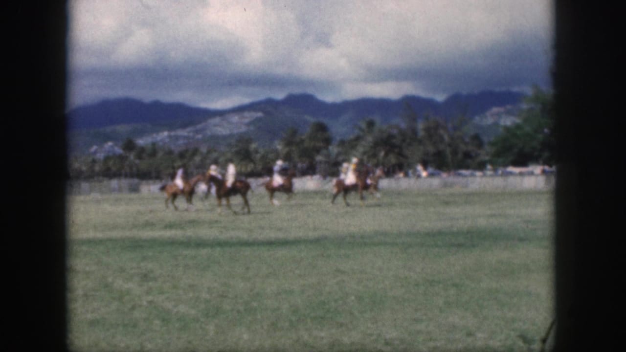1959 Oahu 8mm frame showing several polo players riding across a wide green field with palms and mountains beyond.
