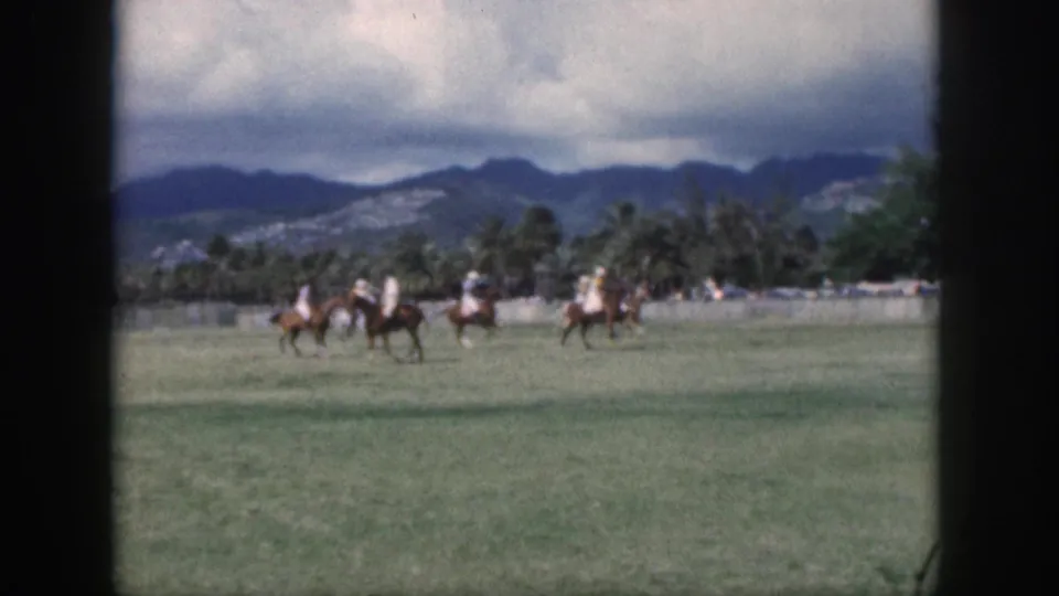1959 Oahu 8mm frame showing several polo players riding across a wide green field with palms and mountains beyond.