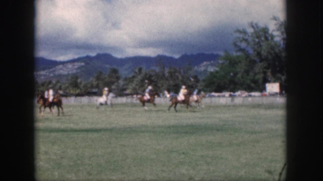 1959 Oahu home-movie frame showing distant polo players on horseback moving across a grassy field under a cloudy sky.