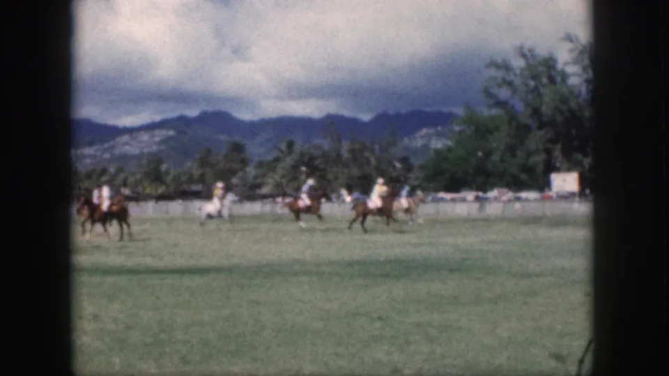 1959 Oahu home-movie frame showing distant polo players on horseback moving across a grassy field under a cloudy sky.